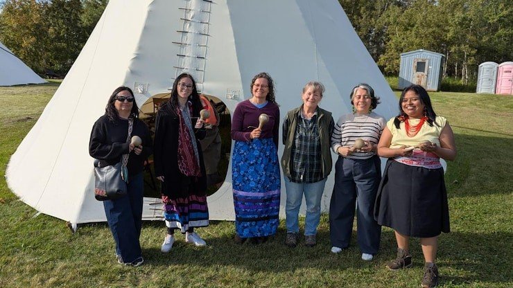 Mujeres representantes de las naciones indígenas Piikani, Tsuut'ina, Cree y Métis de Alberta, de la organización Kairos y Paola Chindoy (a la izquierda) posan frente a un gran tipi blanco, sosteniendo sonajas tradicionales, tras un taller de elaboración de este instrumento.